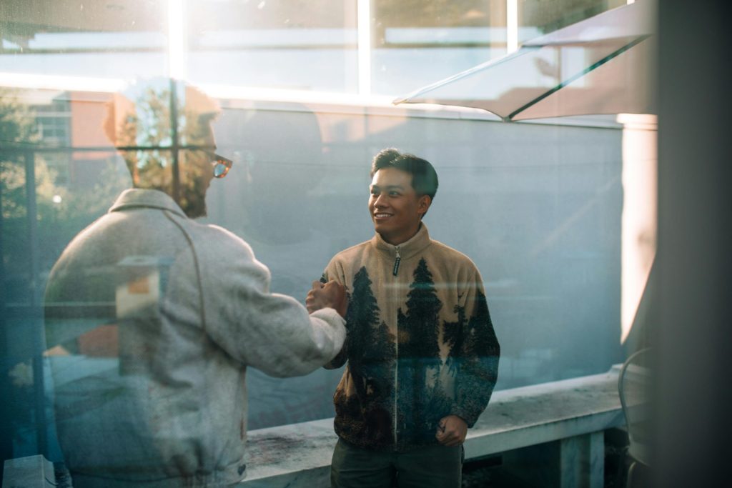Two men smile and shake hands through a glass window, reflecting a warm friendly interaction.